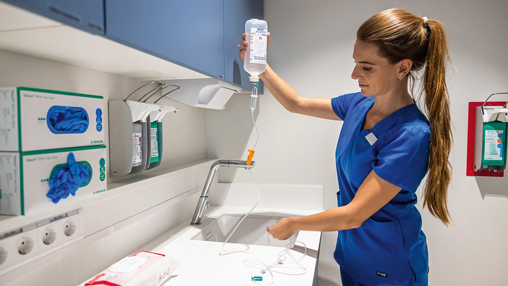 Eine tiermedizinische Fachperson in blauer Arbeitskleidung bereitet eine intravenöse Infusion vor und hält einen Infusionsbeutel sowie Schlauchmaterial über einer klinischen Arbeitsfläche in einem Behandlungsbereich mit medizinischem Zubehör und Geräten.