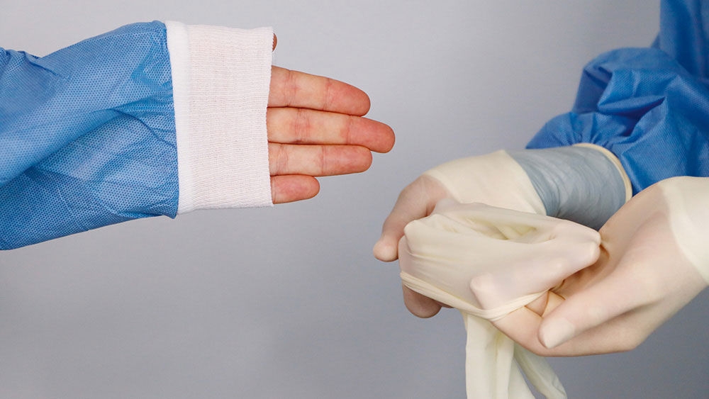 Eine behandschuhte Hand hält einen geöffneten sterilen Handschuh, während eine andere Person in einem blauen OP-Kittel ihre Hand ausstreckt, um ihn anzuziehen, auf einer medizinischen Arbeitsfläche.