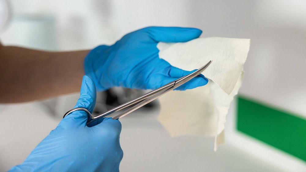 Eine veterinärmedizinische Fachkraft trägt Vasco Nitril-Handschuhe und schneidet ein Stück weiße Gaze oder Vliesmaterial mit einer AESCULAP-Operationsschere.