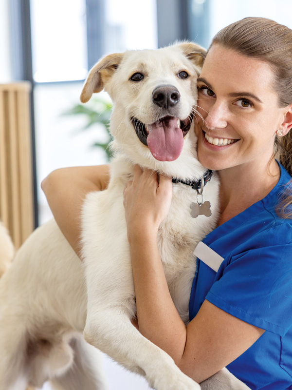 Eine tiermedizinische Fachangestellte in blauer Arbeitskleidung hält einen fröhlichen, hellfarbenen Hund, der in Richtung Kamera schaut.