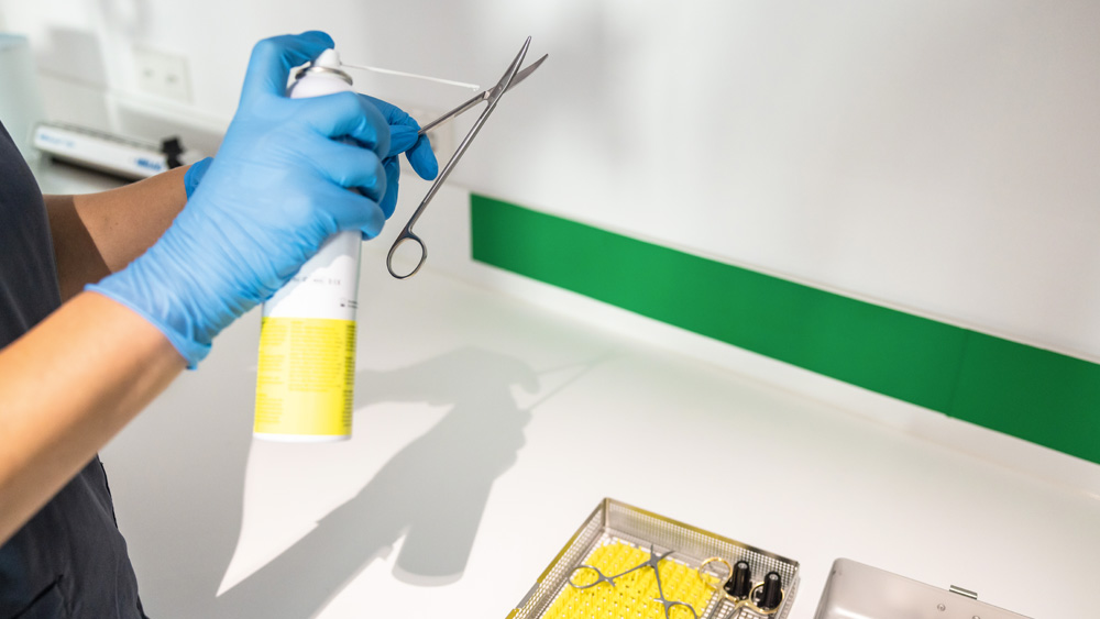 Behandschuhte Hände halten chirurgische Scheren, während in einem Bereich zur Instrumentenaufbereitung Desinfektionsspray aufgetragen wird.
